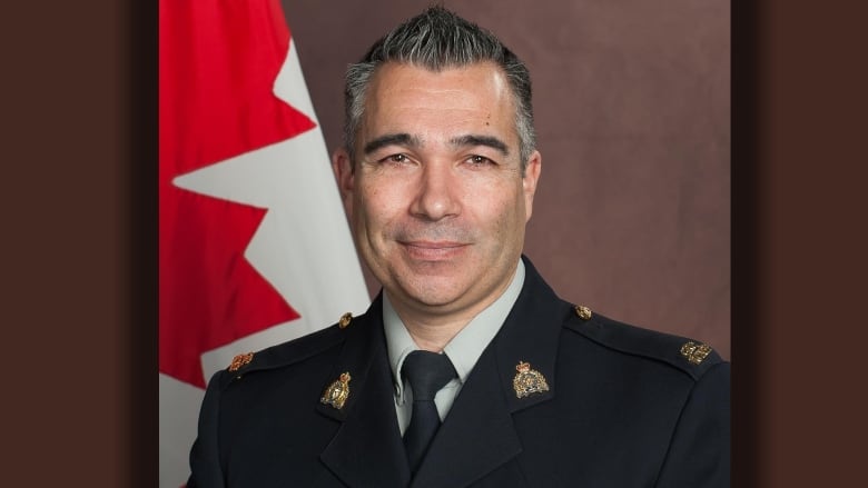 A portrait of a man with short grey hair with a Canadian flag behind him dressed in an RCMP officer's uniform.
