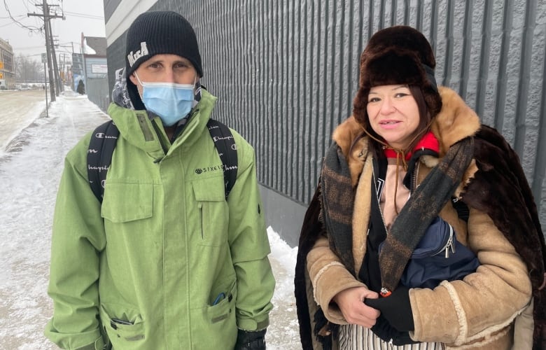 A man in a light green jacket, black toque and blue medical face mask stands outside on a snowy sidewalk next to a woman wearing a brown coat and a fur hat.