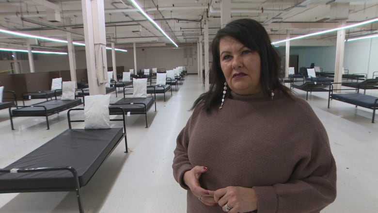 A woman stands in a large room at a homeless shelter. Several empty single beds are seen in rows behind her.