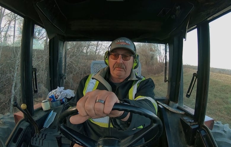 A man with a safety vest, ball cap and safety earmuffs sits in the cab of a tractor with his left hand on the wheel.
