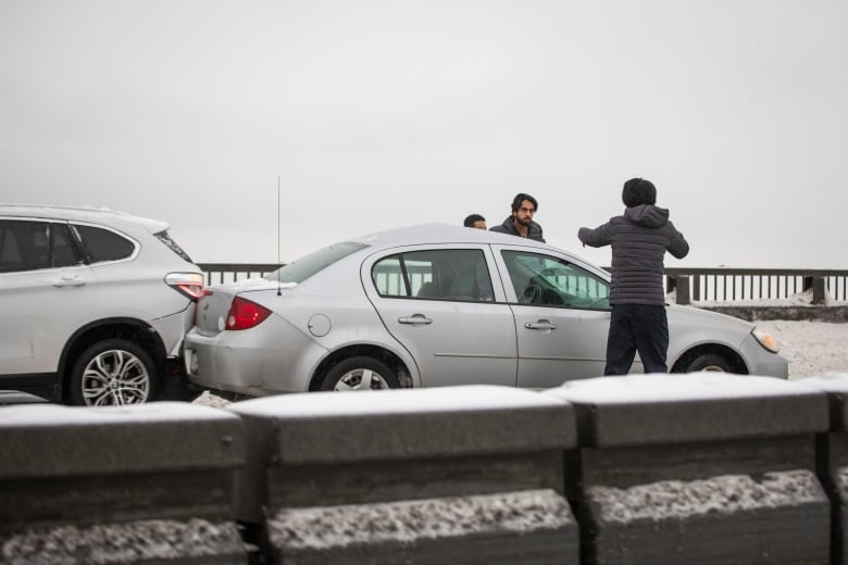 Two men with cars touching each other on a bridge point at each other, amid snowy conditions.