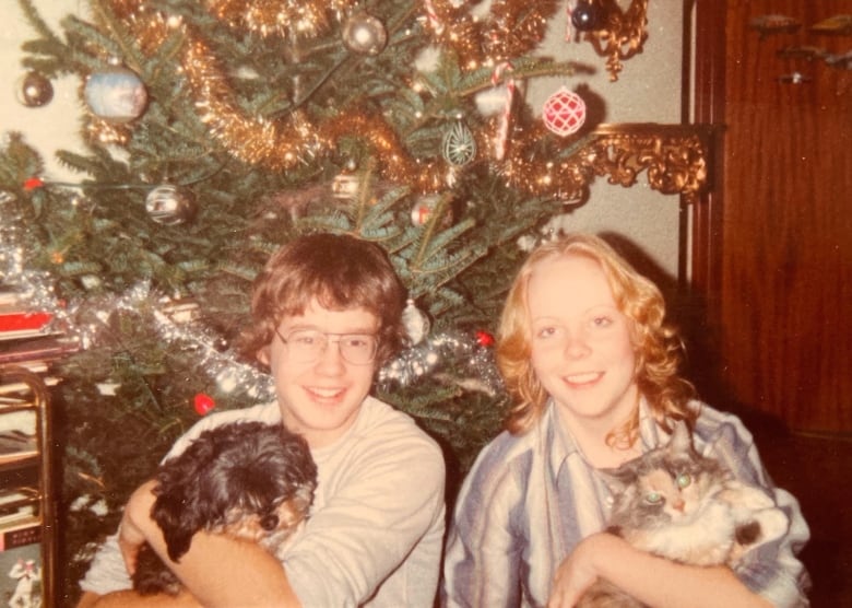 A girl holding a cat and a boy holding a dog sit in front of a Christmas tree. 