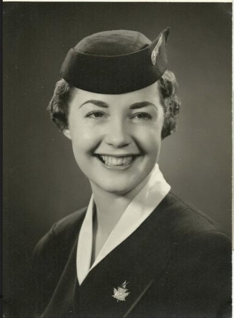 A black-and-white portrait of a young white woman smiling in a midcentury airline attendant uniform. She is wearing a beret-style hat and a blouse under a collared overcoat. 