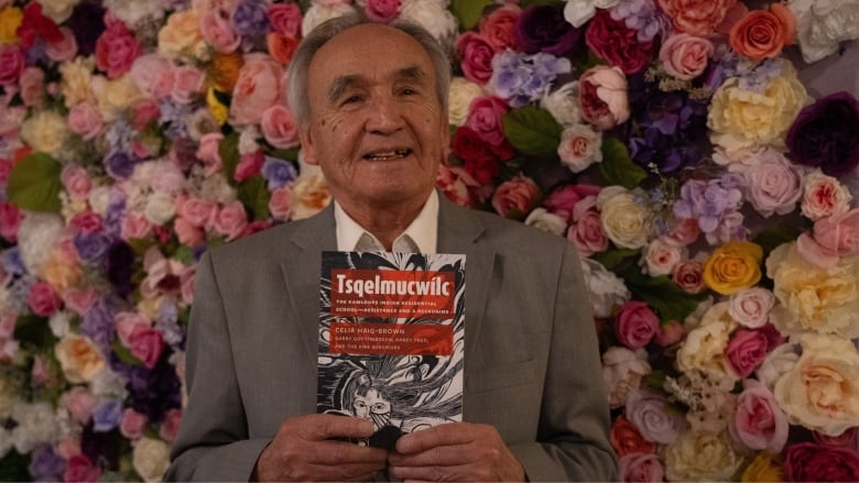 A man smiles holding a book with a flower wall behind him