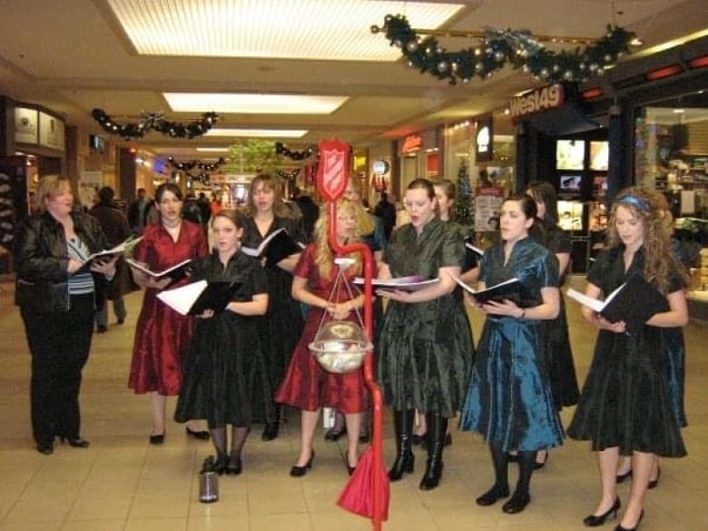 Un groupe de femmes chante dans un centre commercial avec une bouilloire de l'Armée du Salut devant elles.