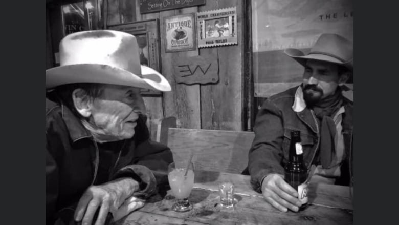 Une photo en noir et blanc montre l'icône de la musique country Ian Tyson penché au-dessus d'une table en train de parler à un autre client.  Tyson porte une veste sombre et un chapeau de cowboy blanc. 
