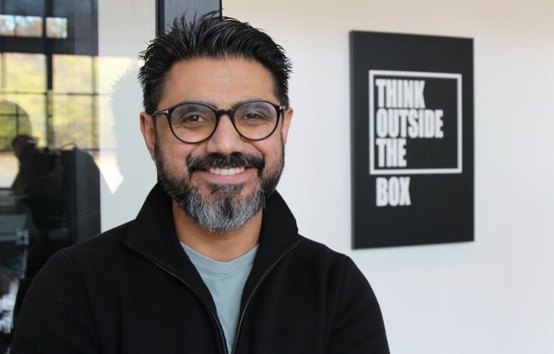 A man in black plastic-rimmed glasses faces the camera, next to a sign that says "Think outside the box"