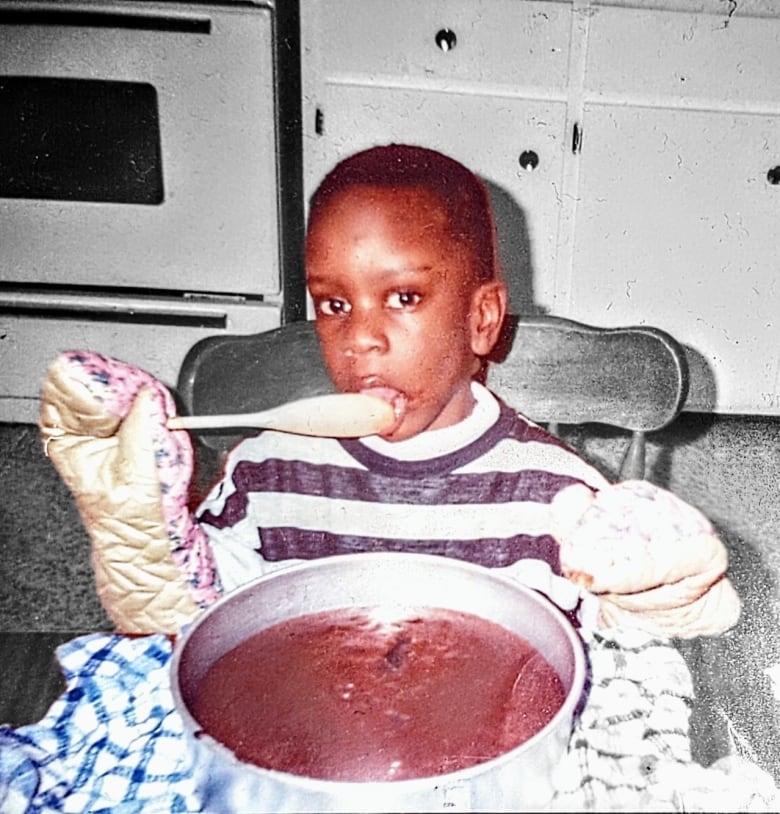 A child sitting at a table, licking a spoon with a chocolate cake in front of him.