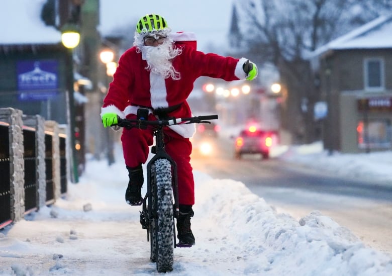 A man dressed as Santa Claus rides a bike through the snow while giving a thumbs up.