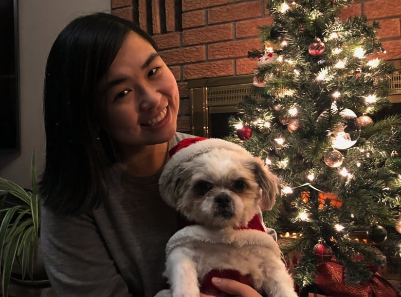 Smiling woman poses in front of her sparkling christmas tree with her shih-tzu dog in her hands. The dog is wearing a santa costume. He does not look happy.