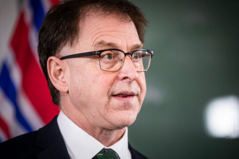 A close-up shot of a man in glasses in mid speech with a B.C. flag in the blurred out background.