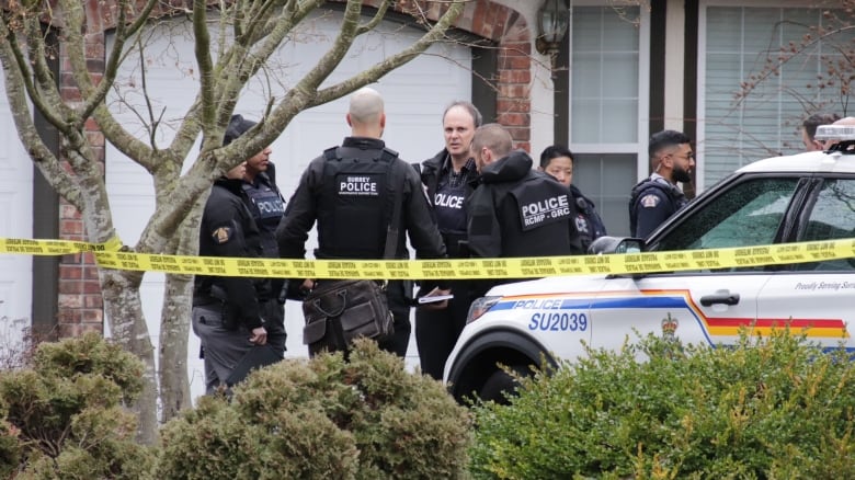 A group of Surrey RCMP officers stand outside of a home where three bodies were found.