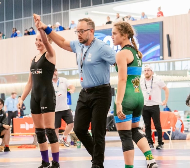 A referee in a gym stands between two teenaged girls in wrestling uniforms, holding up the arm of one of them who's wearing a Team Yukon outfit. 