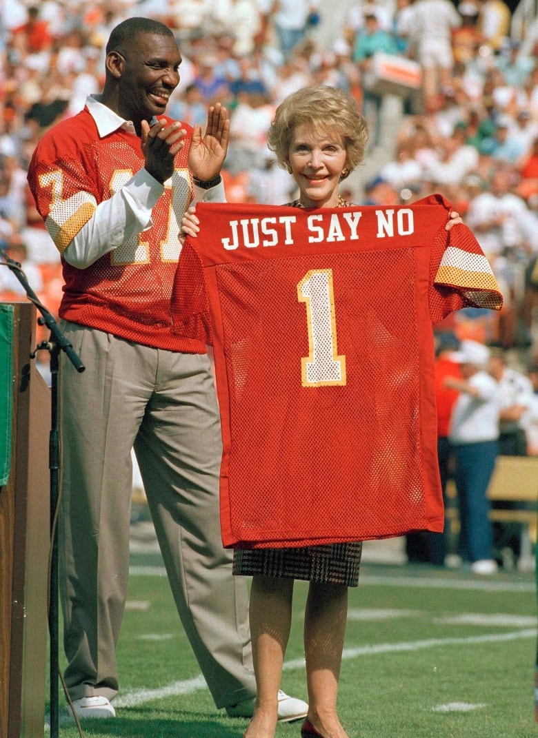 A woman in a sports stadium holds up a jersey with the name 'Just Say No', as a man cheers behind her.