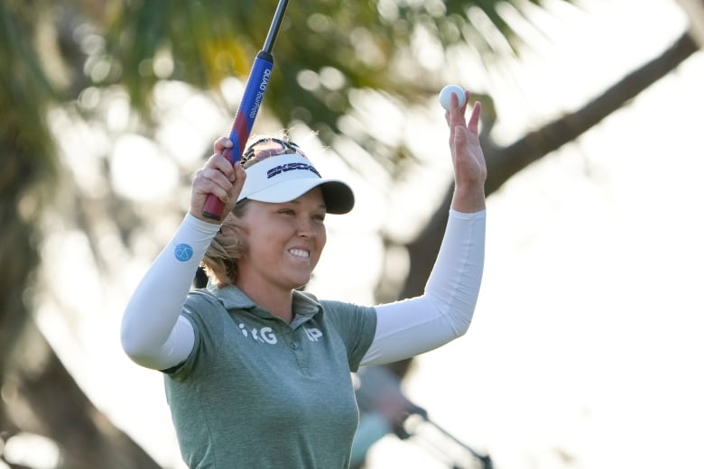 A female golfer smiles while raising her club in her right hand and a ball in her left.