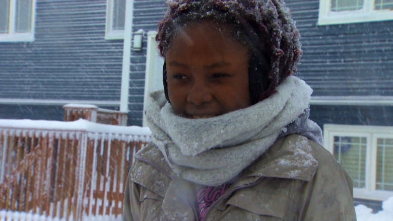 A woman wearing a beige coat and grey scarf stands in her snowy driveway.