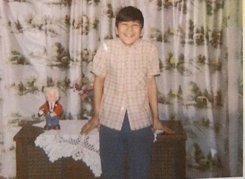 A 1960s photo of a teen boy standing in front of furniture and a curtain.