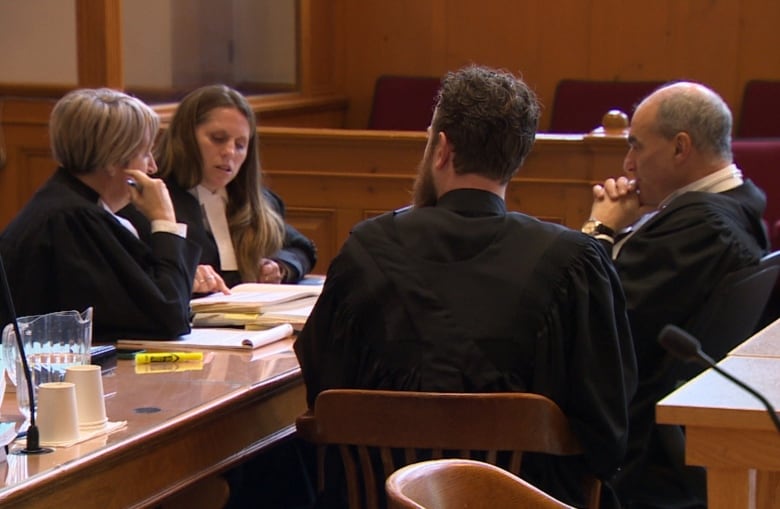 Four people in robes in court sit across a table from each other. One of them is pointing at a document and appears to be discussing it.