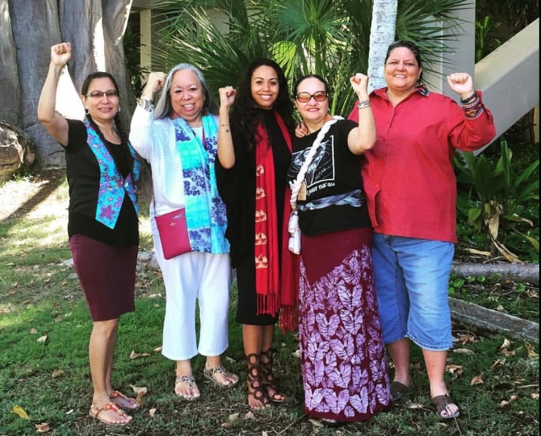 Five women of varying ages stand in front of palm leaves, holding their fists up in the air.