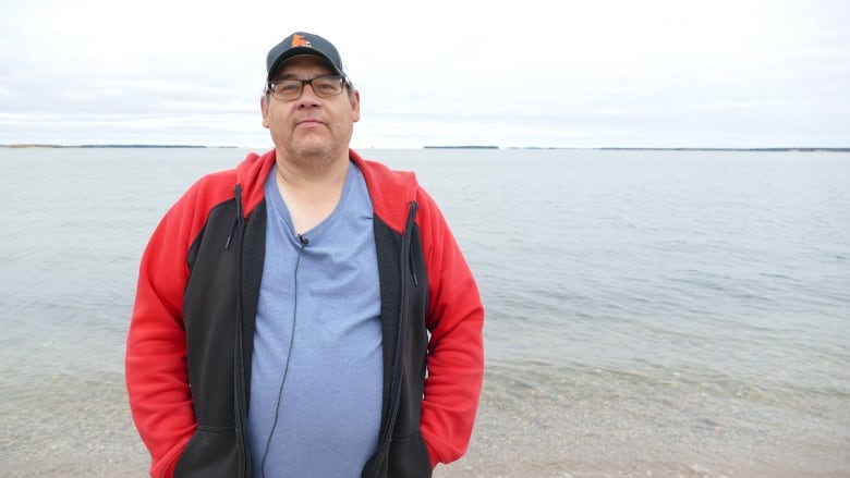 An Indigenous man wearing a cap and glasses stands in front a body of water.