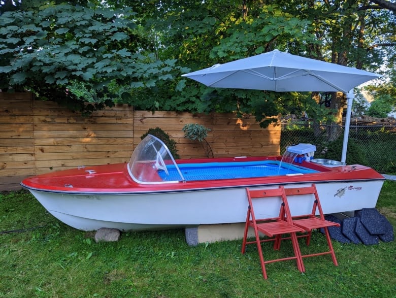 A cherry red motorboat with a pool in the cockpit is seen with a white umbrella hanging over it. In front of it, are two red folding chairs.
