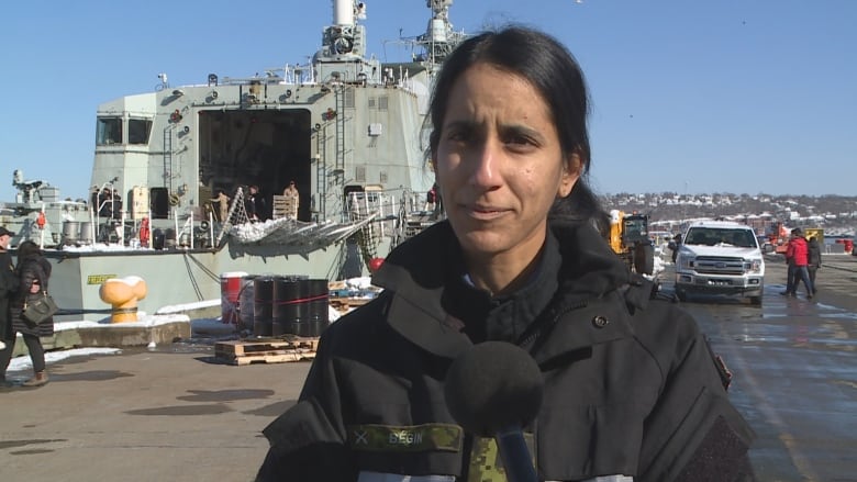 A woman in a black jacket looks at the camera with a frigate in the background. 