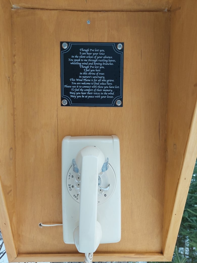 A white vintage, rotary dial phone is affixed to a wooden, booth-like structure which is located outdoors. Above the phone is a plaque that explains its purpose.