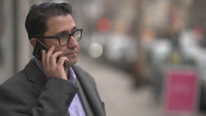 A man with dark hair, wearing black-rimmed glasses and a grey tweed jacket, talks on his cellphone.