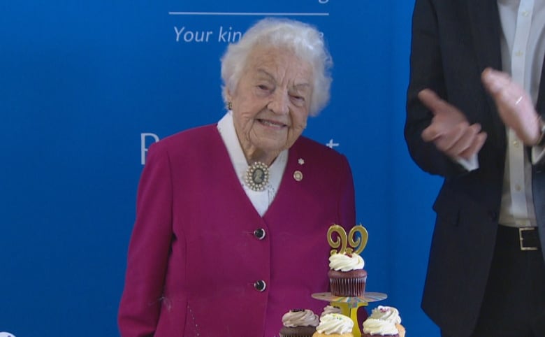 An elderly woman smiles near a pile of cupcakes.