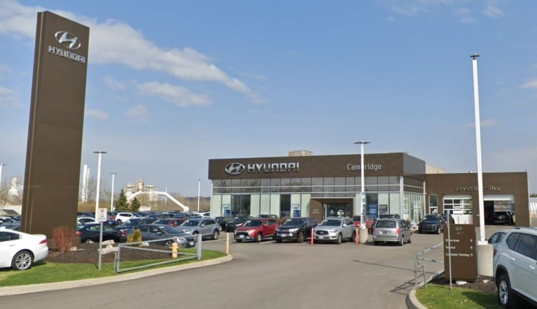A wide-angle photograph of a glass-fronted industrial building that says "Hyundai Cambridge" in large letters on the front. There are about twelve vehicles parked in the lot in front of the building and the sky is almost cloudless.