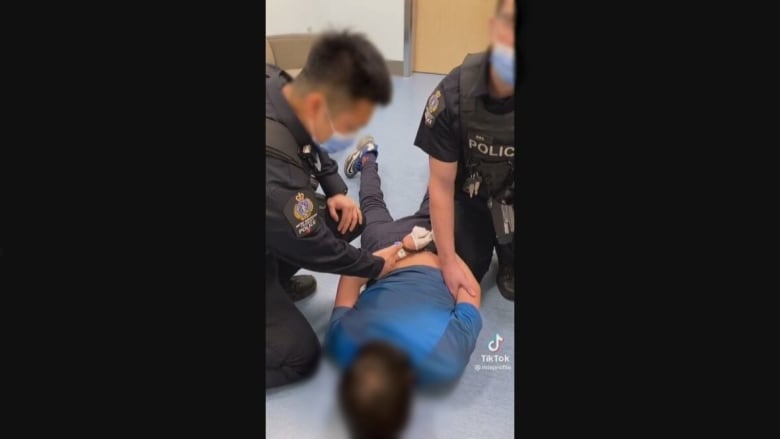 Two officers in uniform hold down a young boy in a blue t-shirt, who is handcuffed and lying on the floor face down.