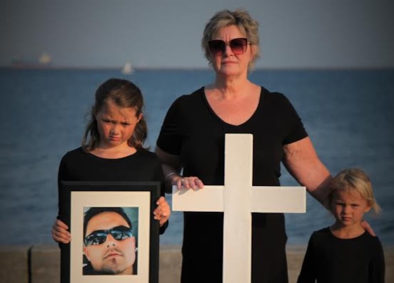 A woman stands with a large white cross and two young girls.