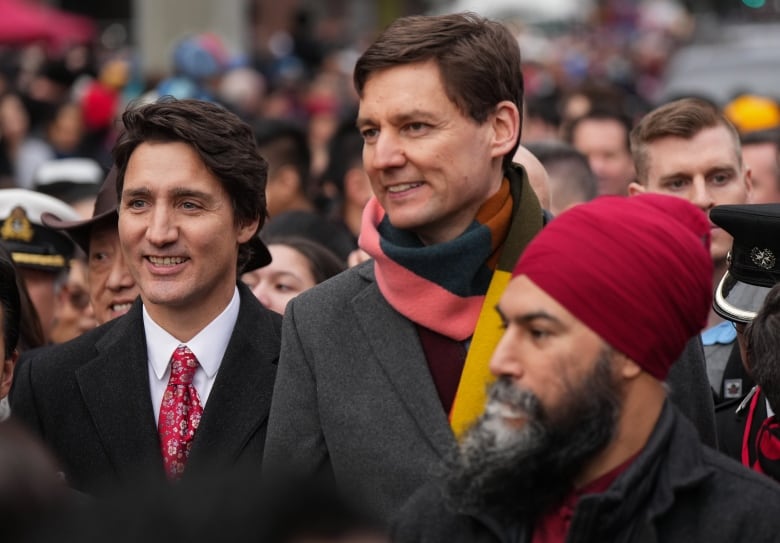 Two white men smile at the head of a parade of people, with a Sikh man off to their left.