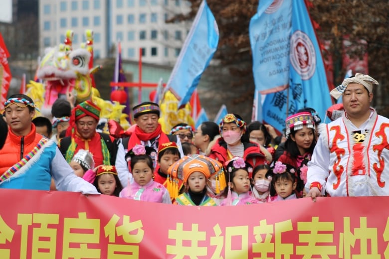 A group of people wearing traditional clothes at the front of a Lunar New Year parade, with a dragon and blue flags visible behind them.