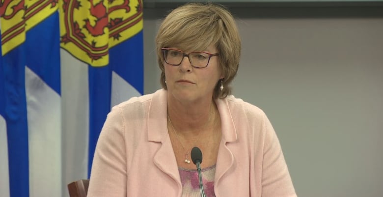 A woman sits at a desk in front of a microphone. Two Nova Scotia flags hang in the background.