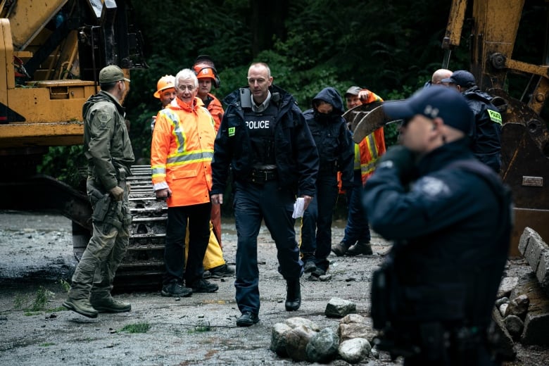 A group of people in high-visibility vests and police officers walk on muddy ground in a forest.