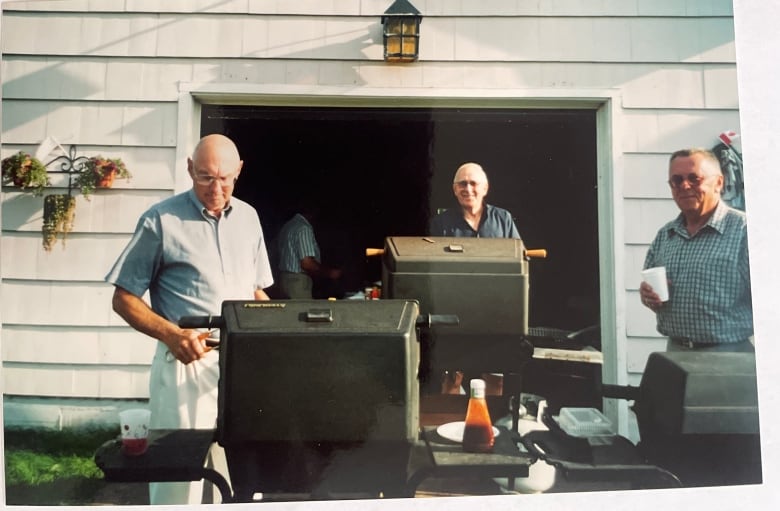 Three men each stand behind a barbecue, two are looking at the camera and one has tongs in his hand and is looking at the grill. Behind them, a white building with an open garage door shows more people are inside.