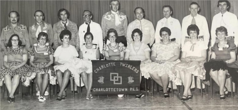 A black and white photo of nine men standing in a row behind nine women who sit on chairs. The women are wearing 1970s style flowing dresses and the men shirts and ties. A sign in the middle reads 'The Charlotte Twirlers.'