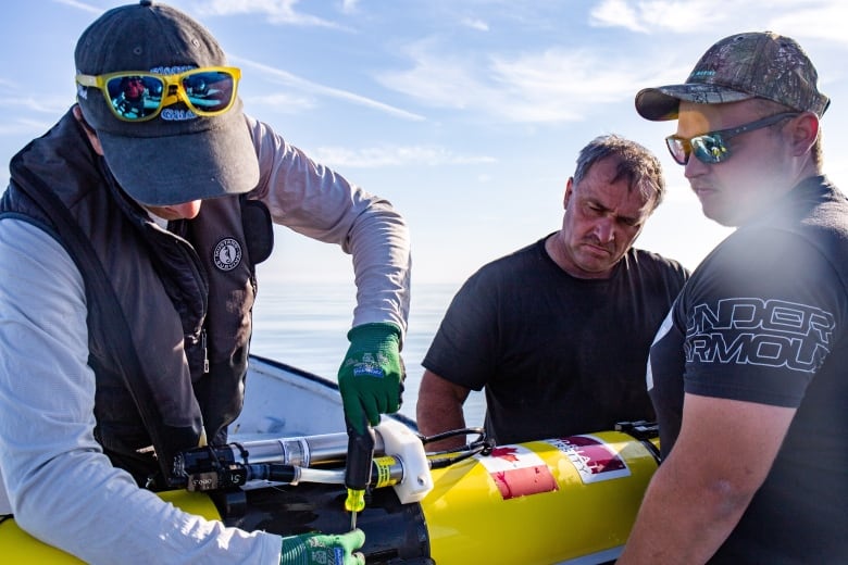 A man uses a screwdriver to fix part of a torpedo shaped drone while two other men look on.