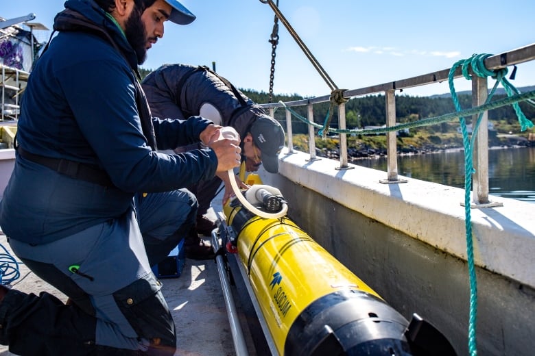 Two men lean over a yellow torpedo-like drone on the deck of a fishing boat.