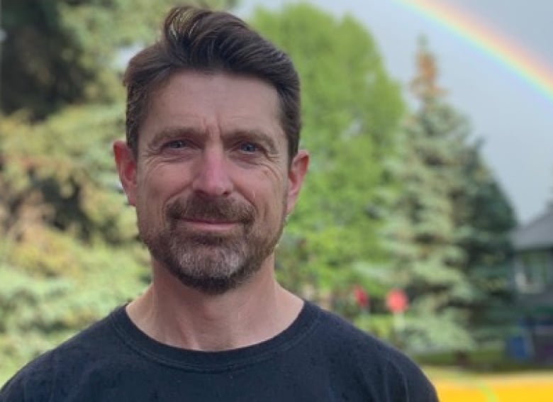 A man wearing a T-shirt looks at the camera with a rainbow in the background.