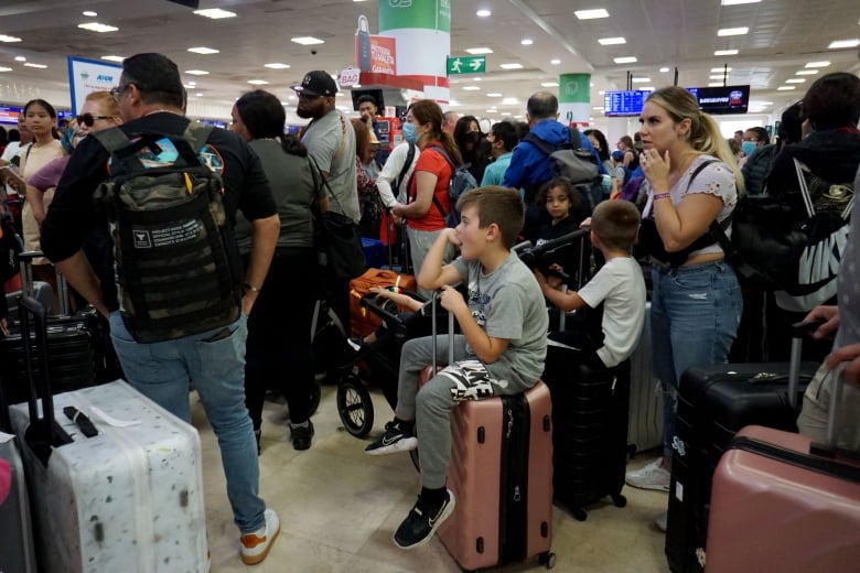 A large number of people, many wearing backpacks, stand around luggage. Some children are sitting on suitcases.
