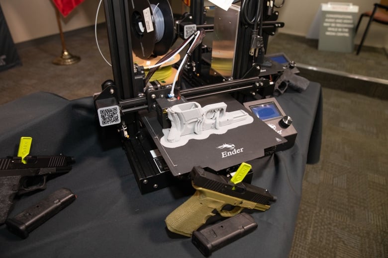 An olive green gun sits on a table with a 3D printer behind it. The 3D printer has a grey firearm on it.