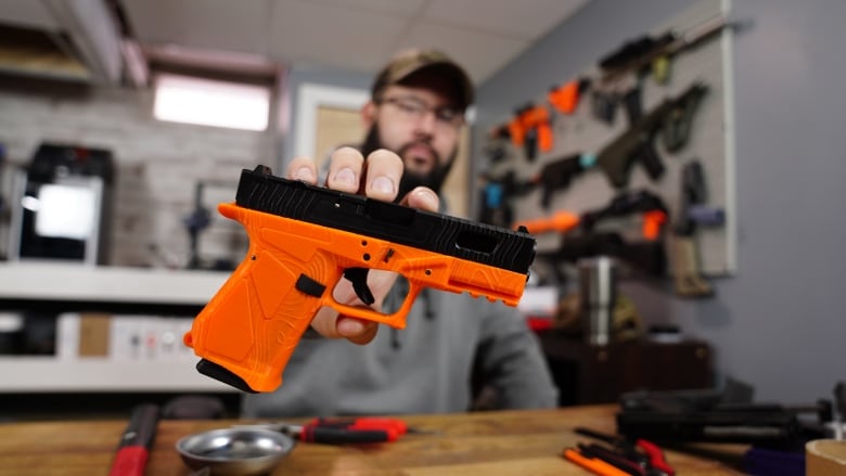 A man holds an orange 3D printed firearm. The firearm is in focus, the man is not.