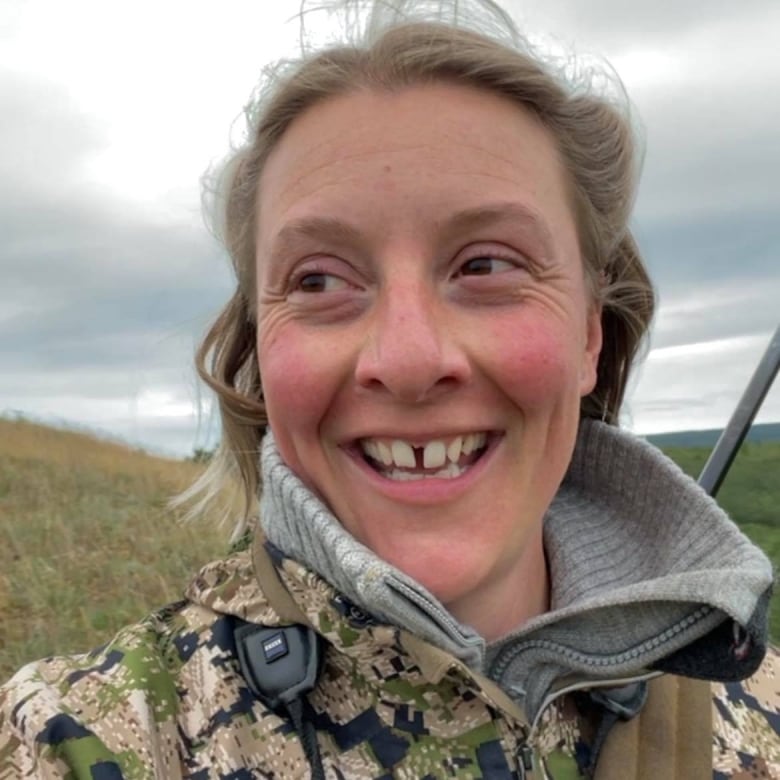 A close-up of a smiling woman's face, standing on a grassy hill with a rifle on her back.