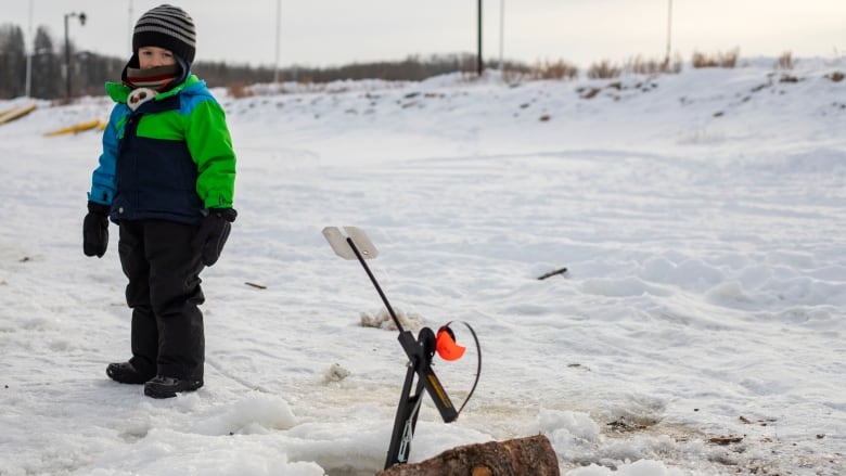A little boy stands by an ice fishing hole outside.
