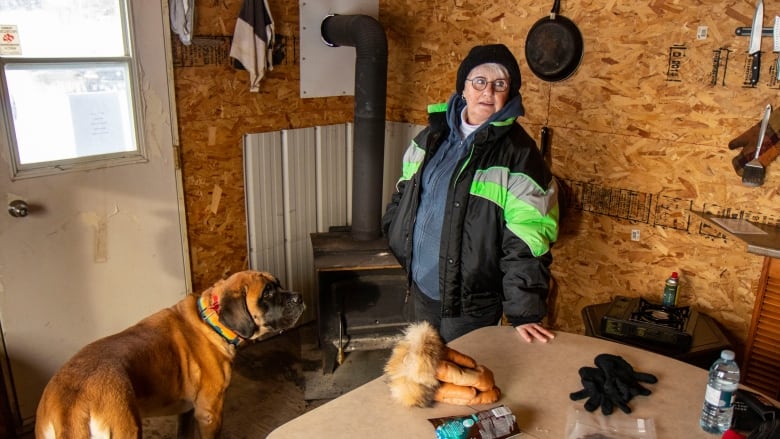 A woman stands in a ice fishing shack with a dog.