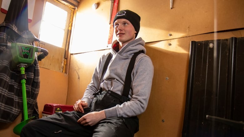 A young man sits in an ice fishing shack.