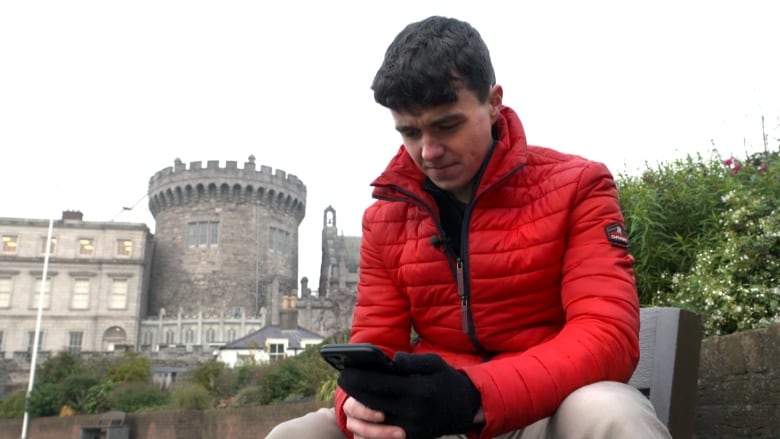 A young man in a bright red jacket on a smartphone in front of a castle.