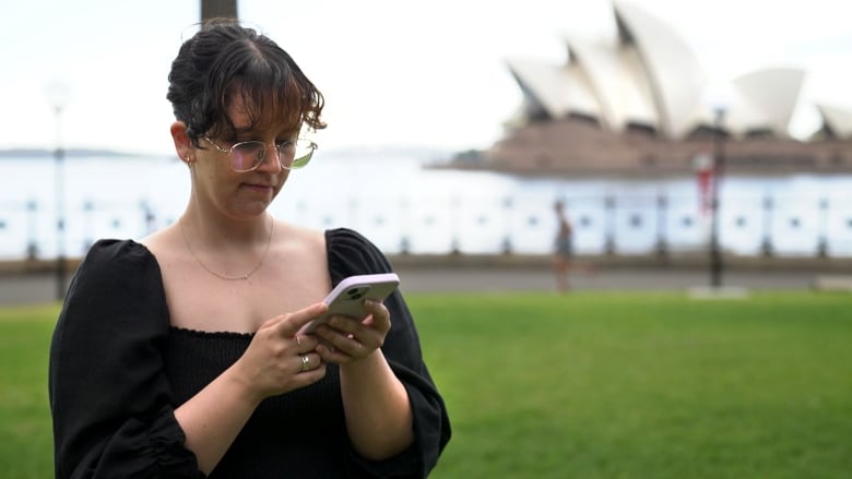 A young woman scrolls on a smartphone in front of the Sydney Opera House. 